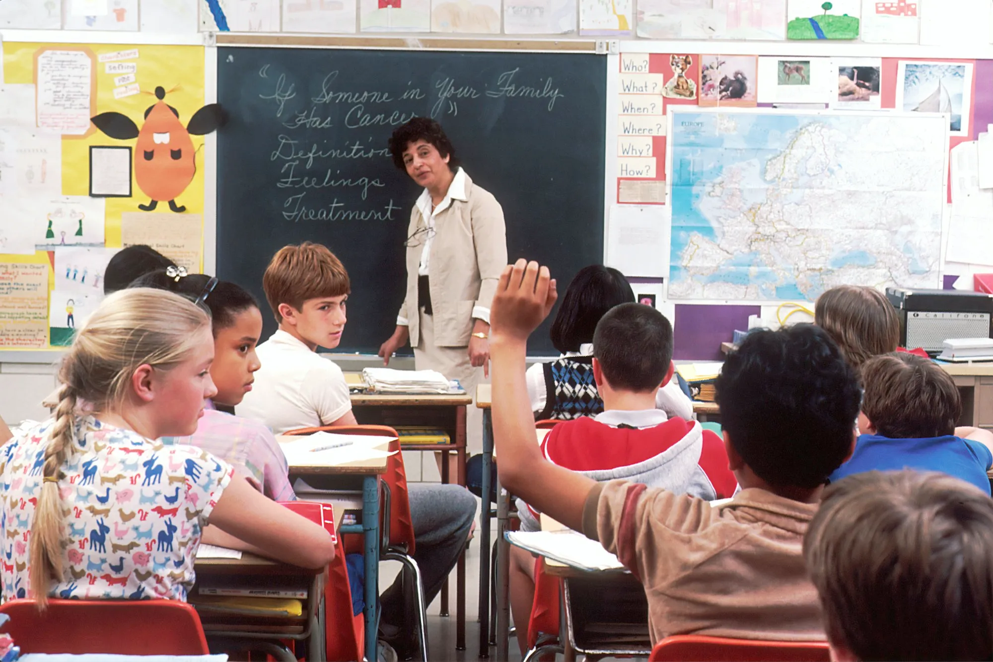 A teacher standing at the front of a classroom by the chalkboard as pupils sit at their desks, one raising a hand.