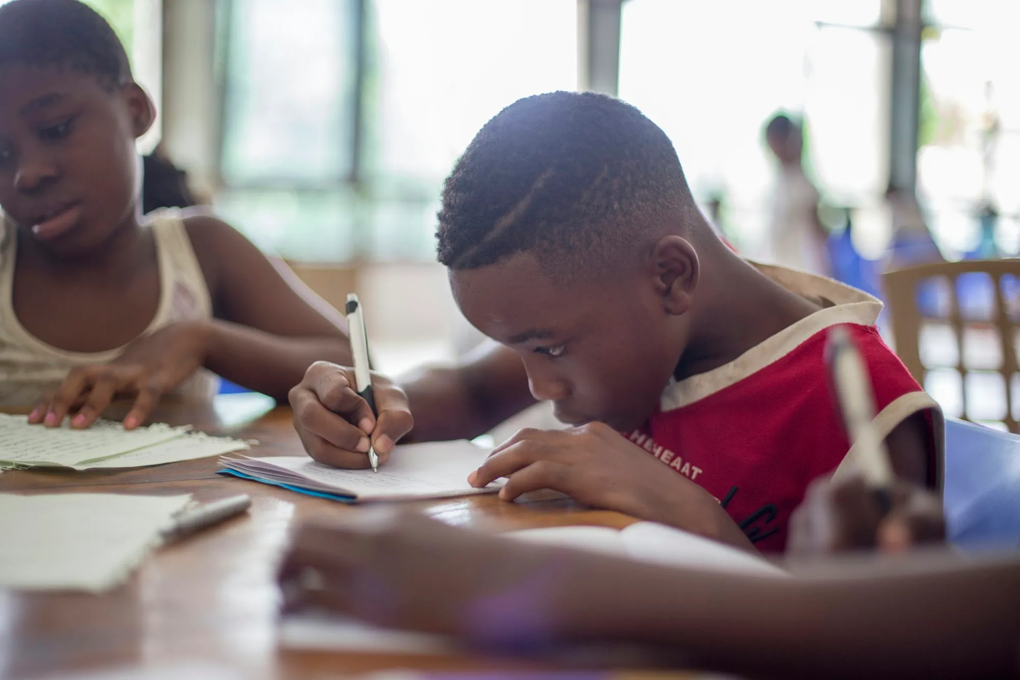 A young boy concentrating as he writes in a notebook, another child studying beside him at a sunlit table.